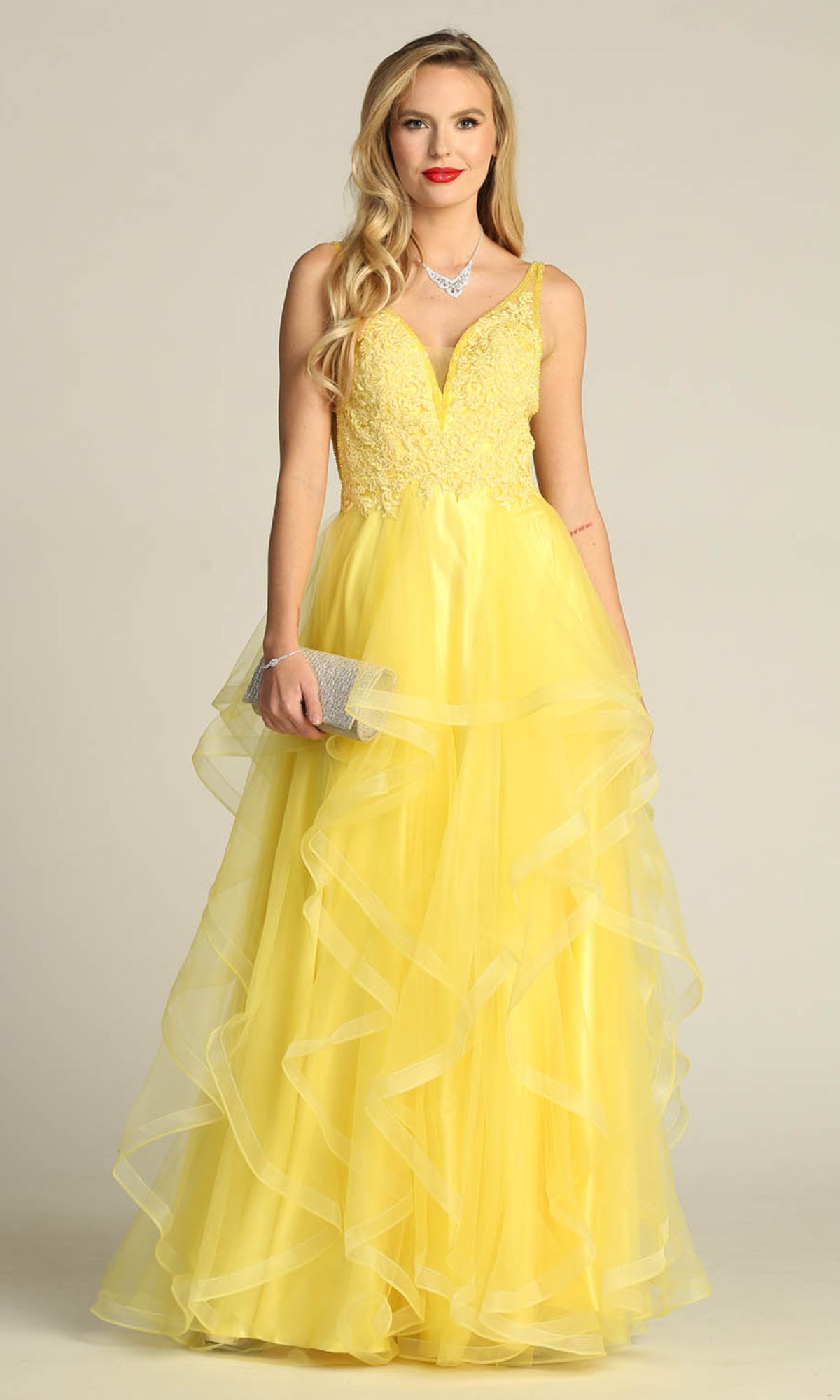 A woman in the Sheer-Sides Long Tiered Prom Dress: Chicas XR6006, sleeveless with a yellow lace bodice and layered tulle skirt, holds a silver clutch and wears a necklace against a plain background.